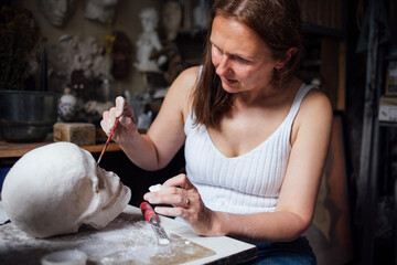 Female artist carefully painting a sculpted head in a creative studio, surrounded by artistic tools and materials, showcasing dedication to the craft of sculpture and artistry
