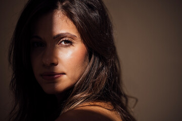Woman with long dark hair gazes thoughtfully, illuminated by soft natural light, showcasing her features and the interplay of shadows and highlights in a serene atmosphere