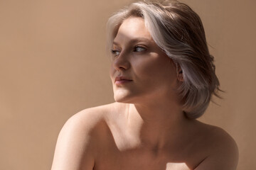 Portrait of a woman with short silver hair, gazing thoughtfully to the side, illuminated by soft natural light against a warm beige background, capturing serene beauty and introspection
