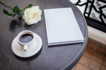 White rose placed beside a blank notebook and a cup of coffee on a round table, creating a serene atmosphere for relaxation and reflection in a cozy setting