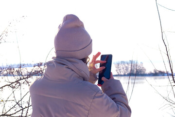 A woman in winter clothes taking a photo of a winter landscape with her mobile phone, showing relaxation and admiration for nature
