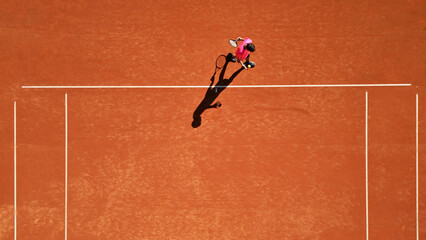 Flat lay photo of a tennis player in a pink shirt practicing on a clay court, focusing on ball handling. Aerial view highlights the red surface and white lines, emphasizing dedication.