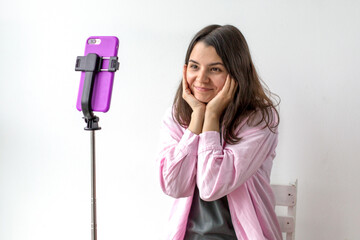 Cheerful young woman in pink shirt holding her face with both hands while smiling at smartphone on tripod, recording video with joy, concept of communication