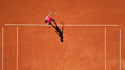 Drone shot of a tennis player in a pink shirt practicing on a clay court, mid-action with a dynamic shadow. Highlights sports training, focus, and athleticism.