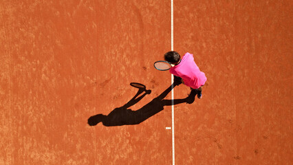 Tennis player in pink shirt practices on clay court, viewed from above. Flat lay photo highlights focused stance and shadow, emphasizing dedication and skill in sports training.