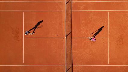 Two tennis players practice on a clay court, shadows stretching across the reddish surface, captured from a bird's-eye view. © stockcopter