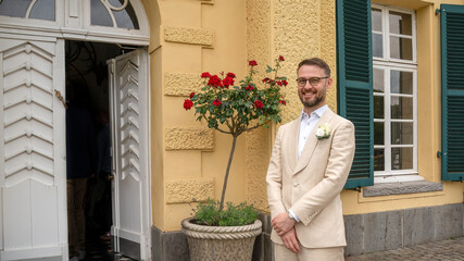 Yowung man in weddding suite waiting for future wife to arrive smiling in front of marriage vacinity