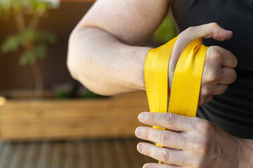 Close-up of muscular man in black shirt wrapping bright yellow strap for wrist strengthening during home armwrestling training for strong forearms. Armwrestling supination exercise with a weight.