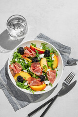 Fresh salad with peaches, arugula, feta, prosciutto and berries in a plate on a light background