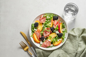 Fresh salad with peaches, arugula, feta, prosciutto and berries in a plate on a light background