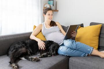 Pregnant woman sitting on couch and reading on tablet with her dog in cozy living room. Happy expectant mother resting on sofa at home. Concept of expectant mother enjoying her pregnancy.