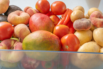 Arrangement of fresh produce arranged inside a modern refrigerator drawer, showing red tomatoes, earthy potatoes, bright bell peppers, and golden nectarines