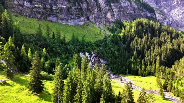 Sibenfalle waterfall cascading down rugged Swiss Alps cliffs into green meadows and evergreen forest beneath a vivid blue sky, a tranquil, unspoiled mountain landscape in Lenk, Switzerland