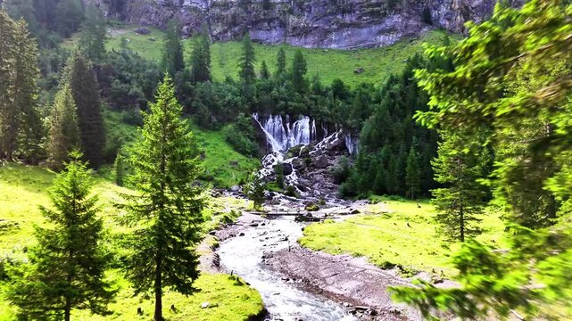 Lenk Sibenfaelle waterfall in Switzerland flows into a rocky river, winding through a green valley with lush conifer trees and a vibrant grassy landscape under a bright sky , drone dolly in shot