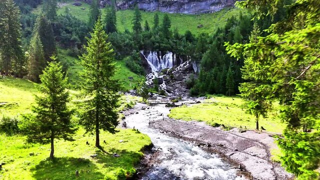 Lenk Sibenfaelle waterfall in Switzerland flows into a rocky river, winding through a green valley with lush conifer trees and a vibrant grassy landscape under a bright sky