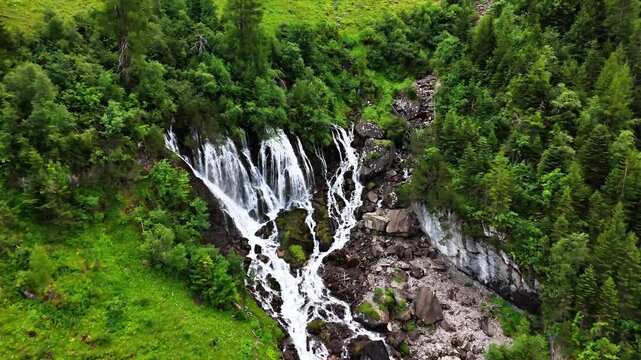 Sibenfaelle waterfall cascading down a rugged mountainside, embraced by thick pine forests and abundant greenery on a overcast day in Lenk, Switzerland, drone push in