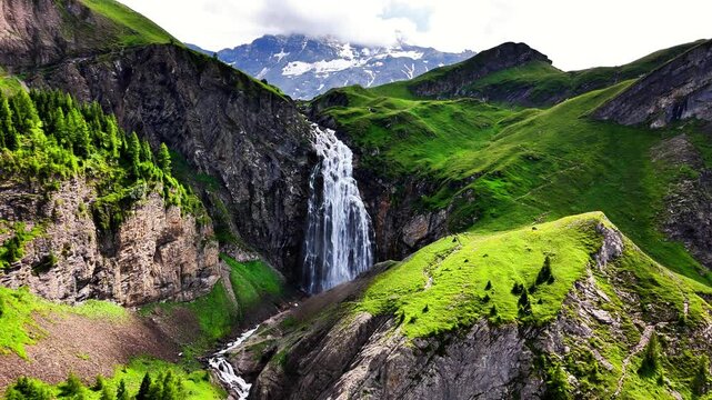 Engstligenfalle waterfall cascades powerfully over rugged cliffs, surrounded by lush green mountain slopes and distant Swiss Alps snow capped peaks, Adelboden, Switzerland, drone static shot