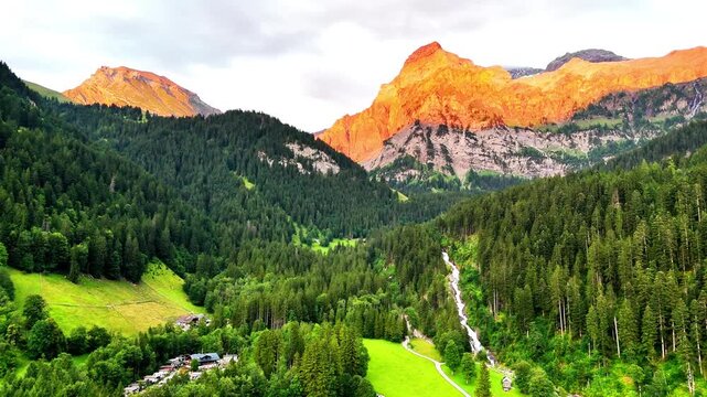 Simmenfalle waterfall flowing from the mountains, surrounded by lush green forests and valleys with traditional buildings, at sunset in Lenk, Switzerland