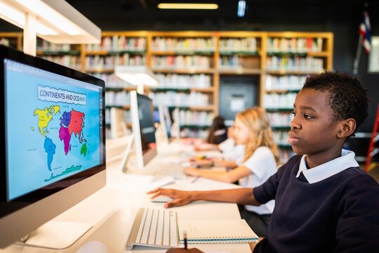 Students in a library using computers for learning. Focused boy at computer, studying maps. Education and technology in a library setting. Elementary school children, African student in library. - Powered by Adobe