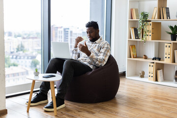 Young adult male sitting on bean bag while using laptop and smartphone in office setting with bookshelf and window view, casual and relaxed atmosphere
