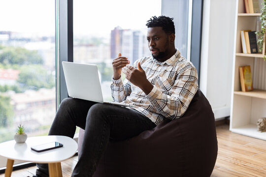 Young adult African American man sitting on bean bag using laptop for video call, communicating confidently in modern room with bookshelf and window view, showcasing work from home
