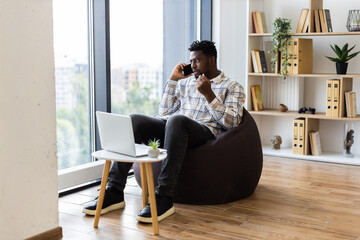 Young adult male sitting on beanbag chair in modern home office multitasking with laptop and phone