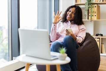 Young adult woman sitting on beanbag holding smartphone gesturing OK sign. Cozy home office environment with laptop, bright window, bookshelf. Concept of confidence work-life balance positivity
