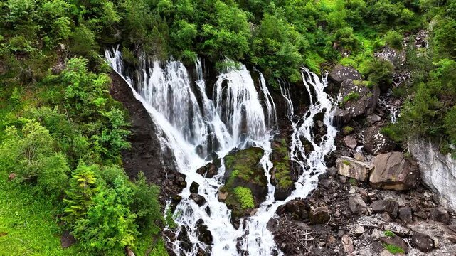 Sibenfalle waterfall showcasing multiple streams of water cascading over dark rocks and mossy terrain, surrounded by trees and representing the power of nature, Lenk, Switzerland, drone static shot