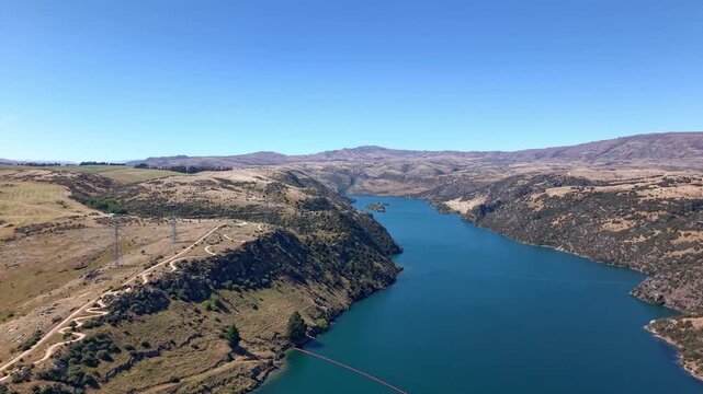 Stunning aerial drone shot flying diagonally to reveal the blue waters and surrounding hills of Lake Roxburgh in Central Otago, New Zealand.