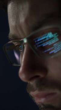 Close up of concentrated programmer in glasses works late at night, lines of code reflecting in his eyeglasses. Software engineer is engaged in coding, data science, or AI development. Vertical video