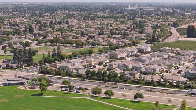 Drone ascending above grassy park toward Ripon, California, showing residential houses, roads with traffic, and trees stretching to the horizon.