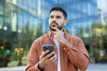 Thoughtful young indian businessman standing outdoors in a city, holding his smartphone and gazing upward in focused contemplation, planning strategy and future success