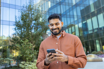 Young handsome indian man smiling while texting on his smartphone outdoors in front of a modern...