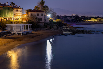 Deserted night beach in Side's old town with illuminated promenade, tiled-roof houses and water reflections under Mediterranean sky. Low season tranquility. Antalya, Turkey.

