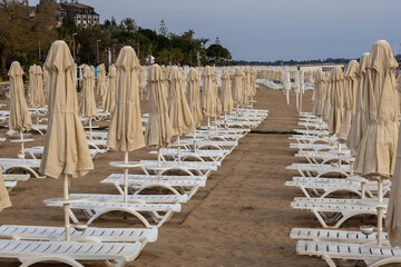 Empty beach with rows of white sunbeds and folded parasols receding into distance under post-sunset light. Palm trees on promenade. Low season tranquility. Kumköy, Antalya, Turkey.