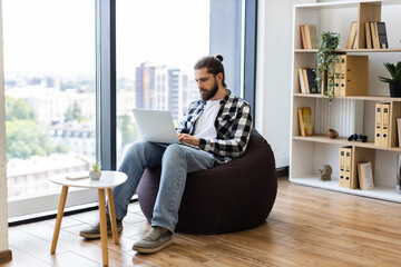Young adult Caucasian male working on laptop comfortably seated on bean bag in modern home office with daylight through large window and organized bookshelf in background..