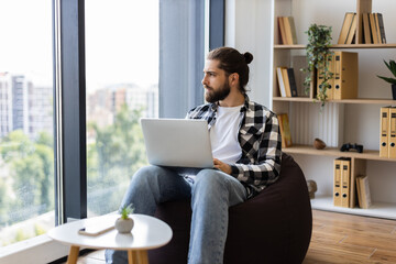 Young adult male with beard and long hair using laptop in cozy home setting. Relaxed atmosphere, comfortable seating, natural light, casual outfit, productive moment, contemporary interior design