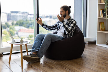 Young adult male sitting in cozy room using smartphone for video call communication