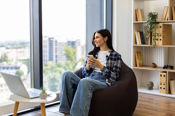 Young adult woman sitting on beanbag chair by window using smartphone. Wearing casual outfit in modern, bright apartment with bookshelves. Relaxed moment of leisure, enjoying serene environment.