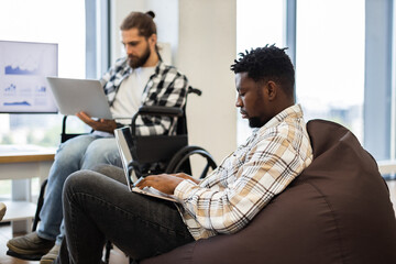 Coworkers, one seated in wheelchair and other on beanbag, working with laptops, showcasing collaboration, inclusivity and technological focus in professional office emphasizing teamwork accessibility.