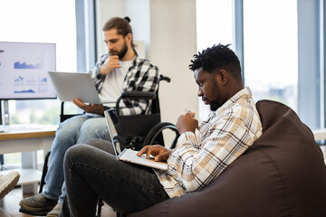 Coworkers, one seated in wheelchair and other on beanbag, working with laptops, showcasing collaboration, inclusivity and technological focus in professional office emphasizing teamwork accessibility.