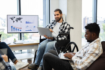 Young adult man in wheelchair presenting in office environment with diverse coworkers using technology displays