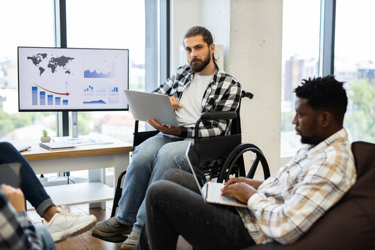 Two young adults engaged in business discussion in wheelchair-accessible environment with digital charts. Focused diverse team working on presentation in office highlighting inclusion and teamwork.