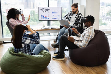 Group of diverse adults using virtual reality headsets and laptops in modern office setting. Individuals of various backgrounds collaborating in technology-driven workspace fostering creativity