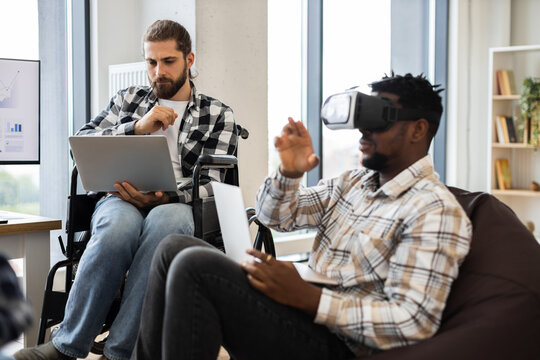 Two adult men, one using virtual reality goggles, collaborate on technology-based projects in office.