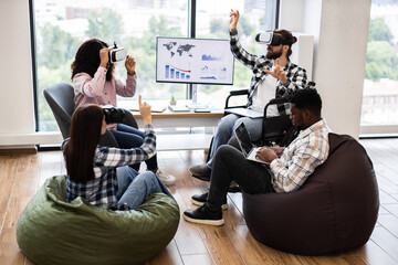 Young adults engage with virtual reality devices and laptops in collaborative office. Diverse teams demonstrating technology interaction seated on floor and chairs creating innovative atmosphere.