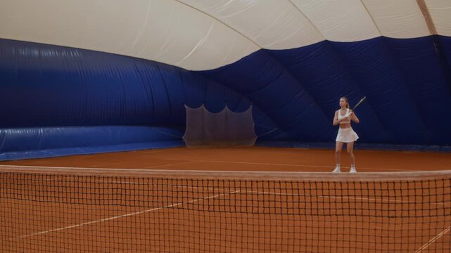 Wide shot of young adult woman in tennis wear hitting ball back over net to opponent practicing lob shot while playing active sports game on indoor red clay court
