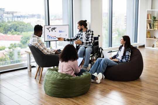 Diverse professionals discussing financial report in office. Includes young wheelchair user leading conversation and mixed gender team interacting around monitor with graphs.