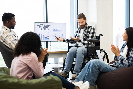 Multiracial team including young adult man in wheelchair collaborating on business presentation, discussing economic trends in open workspace. Evening meeting showcasing teamwork, inclusion
