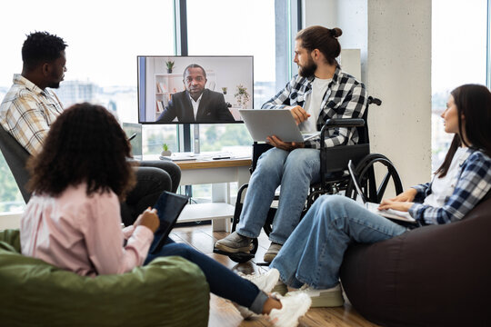 Group of young professionals collaborating in office during video call with male manager. Meeting showcases workspace diversity including individuals of different ethnicities and backgrounds.